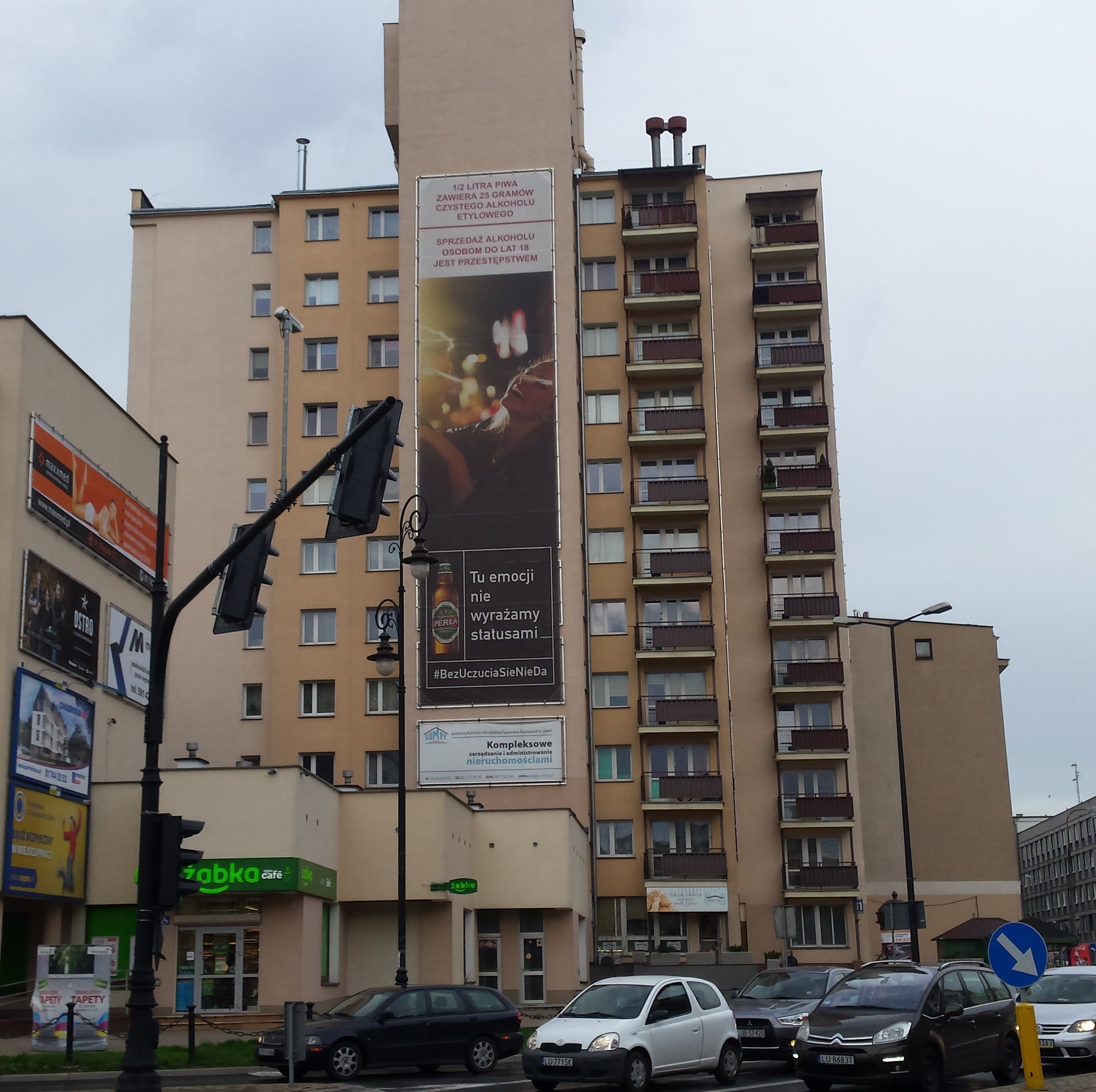 Traffic light and multiple cars in front of a beige multi-story residential building with a large vertical advertisement banner in Polish on the facade.