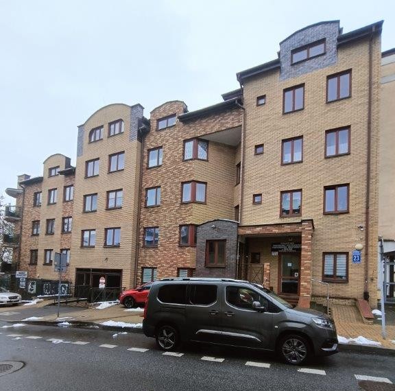 Multi-story residential building with beige brick facade and various window shapes, parked cars in front on a wet street with some snow patches.