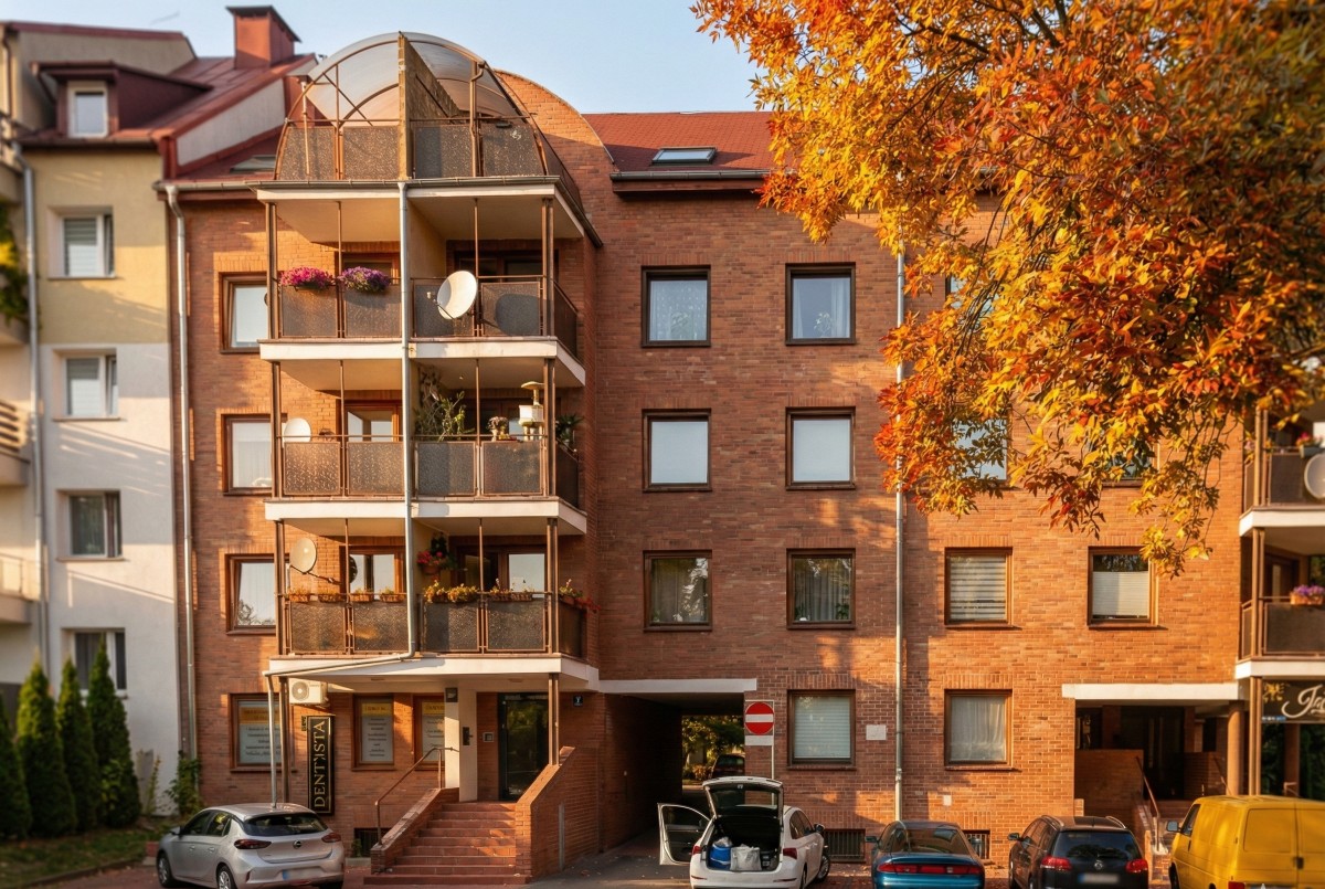 Brick apartment building with balconies, parked cars, and autumn tree with orange leaves in foreground.