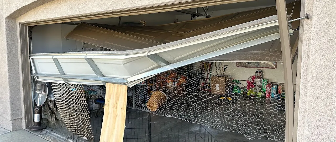 Partially open, damaged garage door bent inward with a wire mesh barrier, showing cluttered items inside the garage.