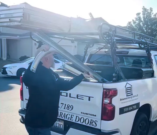 Man unloading metal garage door part from a white pickup truck with ladder rack in a sunny parking lot.