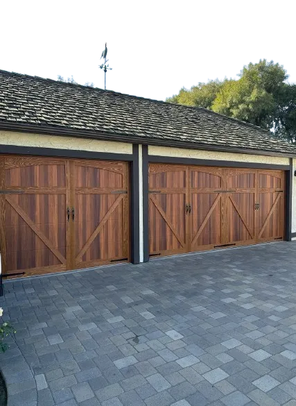 Two closed wooden garage doors with decorative cross patterns, mounted on a house with a shingled roof and paved driveway.