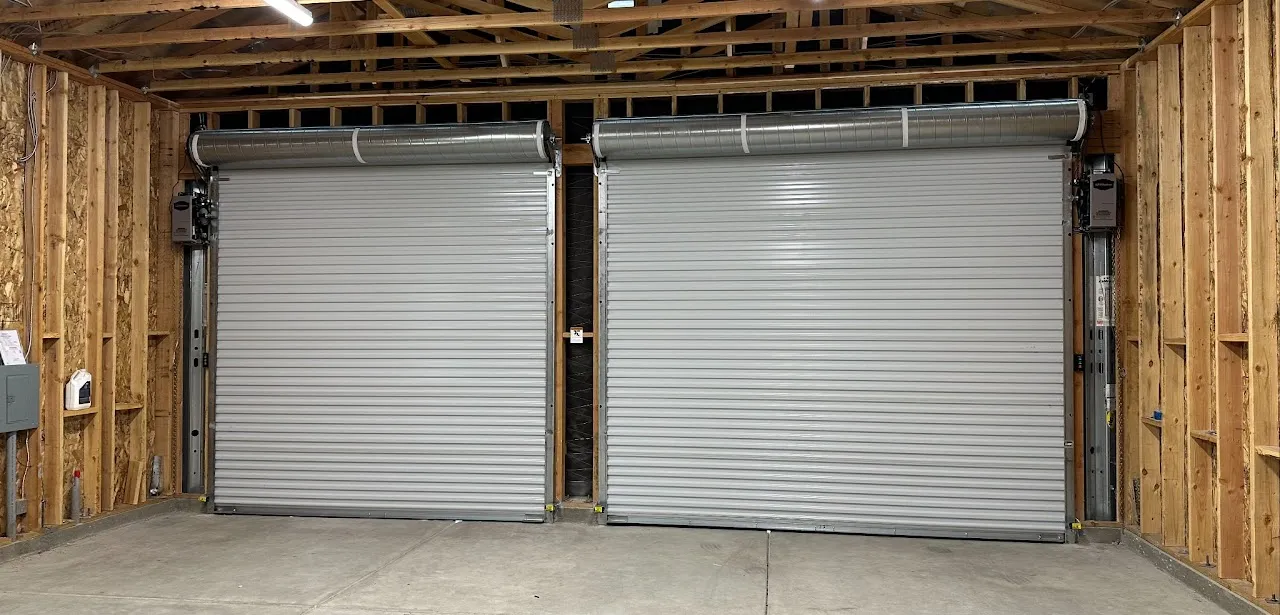 Interior view of a garage or warehouse showing two closed metal roll-up doors framed by unfinished wooden walls and concrete floor.