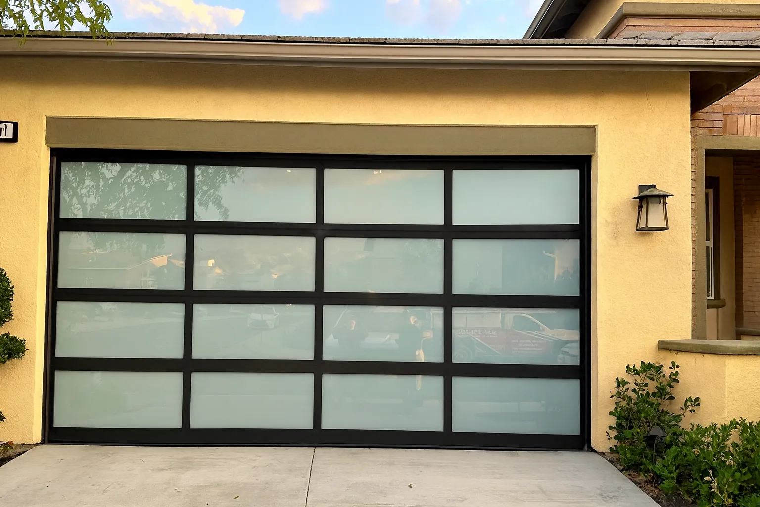 Modern translucent glass garage door with black metal frame on a yellow stucco house exterior.