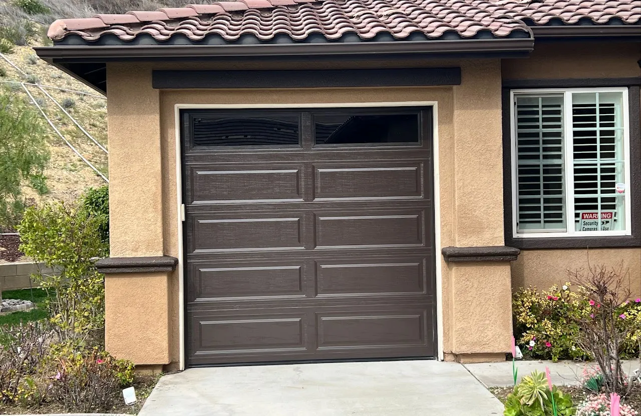 Closed dark brown garage door with windows at the top on a beige stucco house with terracotta roof tiles and nearby plants.