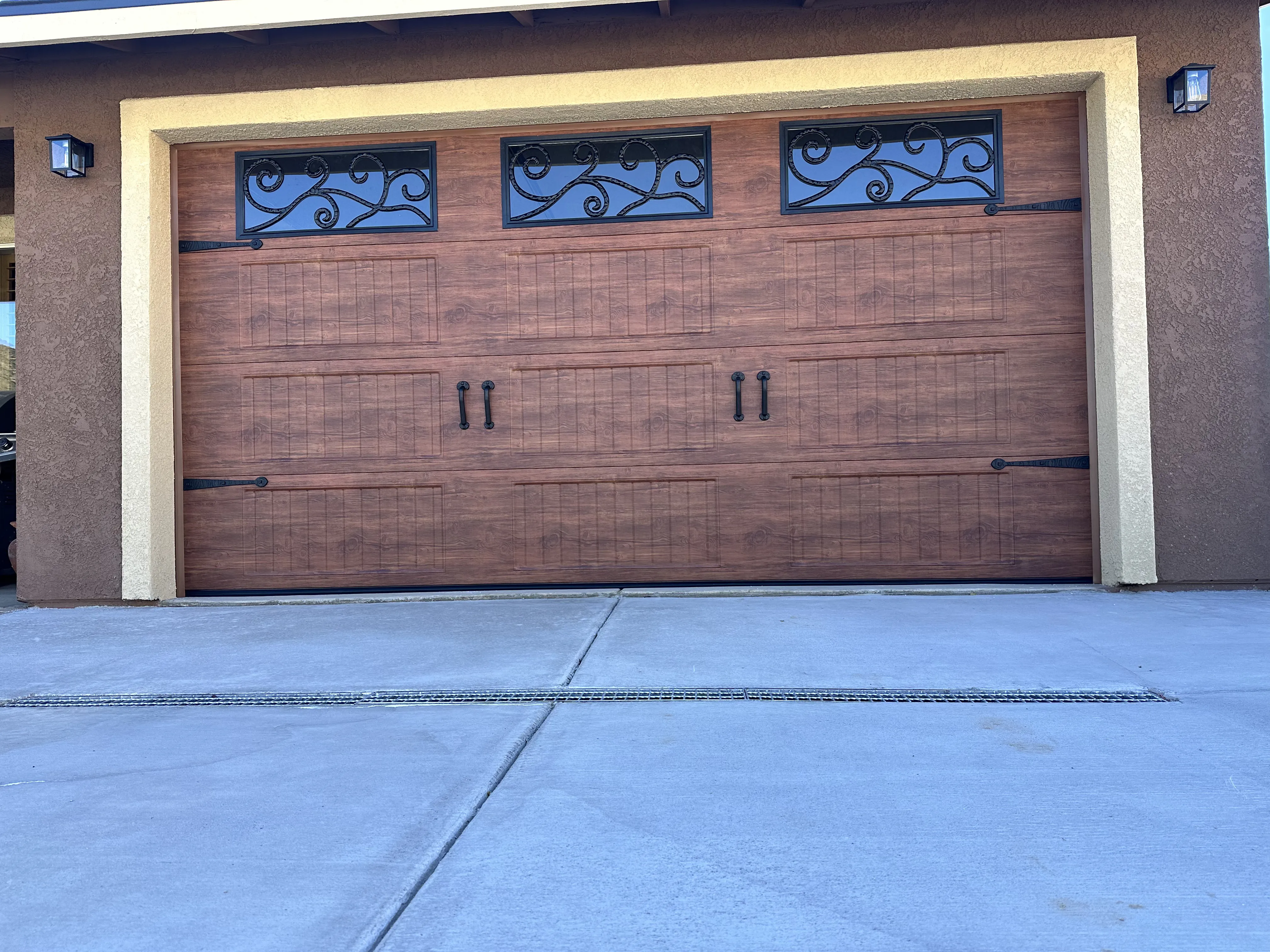 Brown wooden garage door with three decorative windows featuring black wrought iron scrollwork and two sets of black handles.