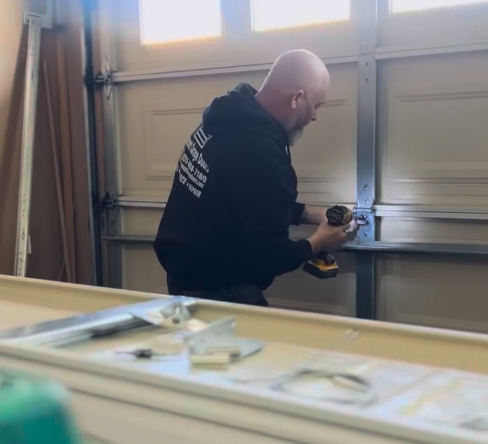 Man using a cordless drill to install hardware on a garage door inside a workshop.