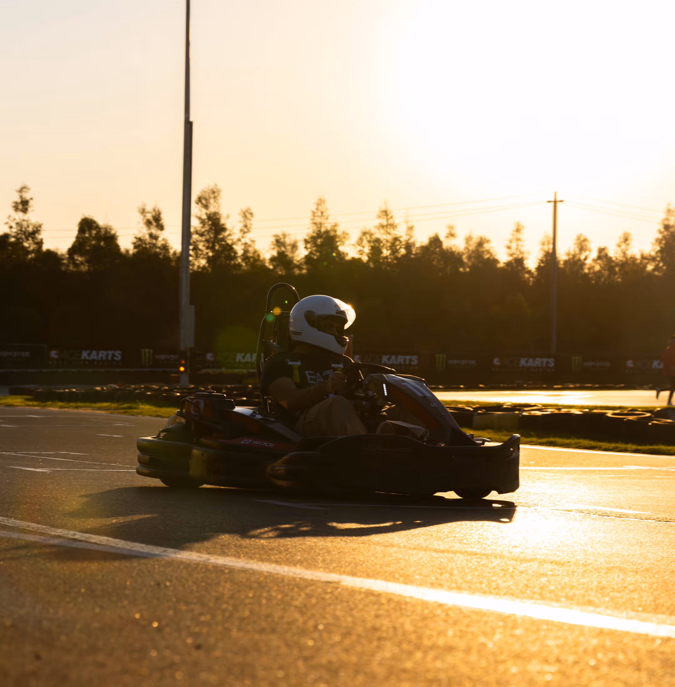 Group of six people in racing helmets with clear visors and MZR logos behind dark textured barrier, one person pointing upward, warm golden lighting