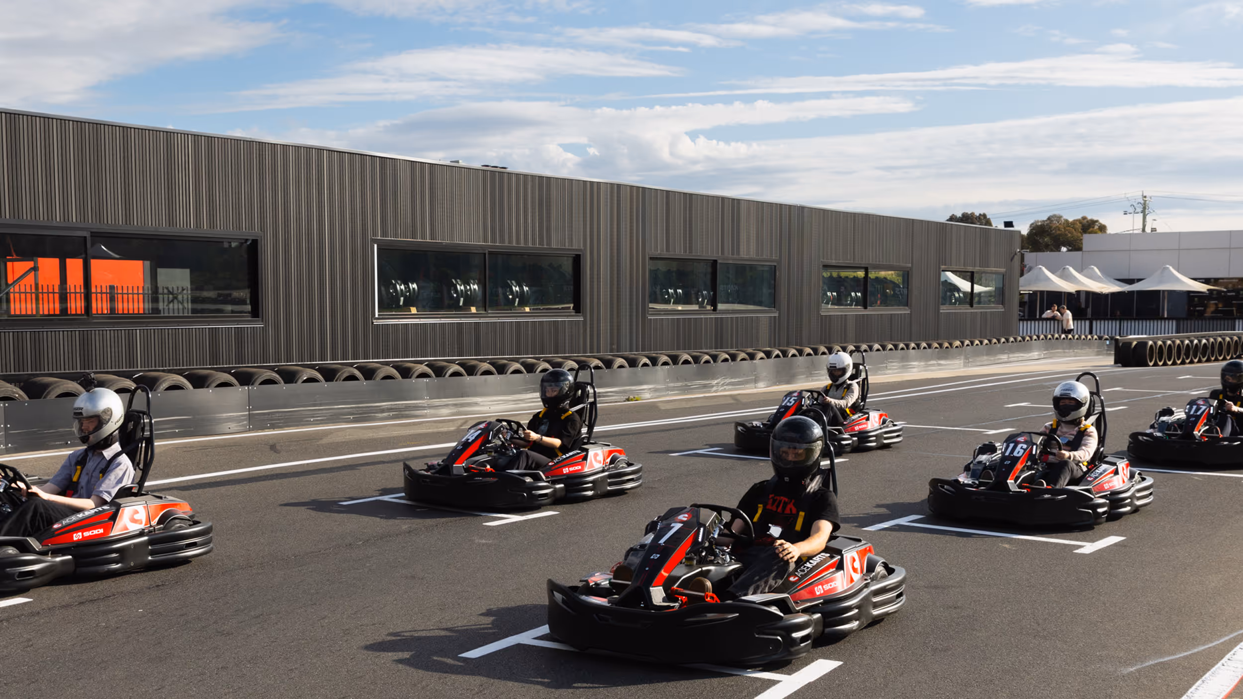 Six individuals wearing helmets driving go-karts on an outdoor race track near a building with large windows.