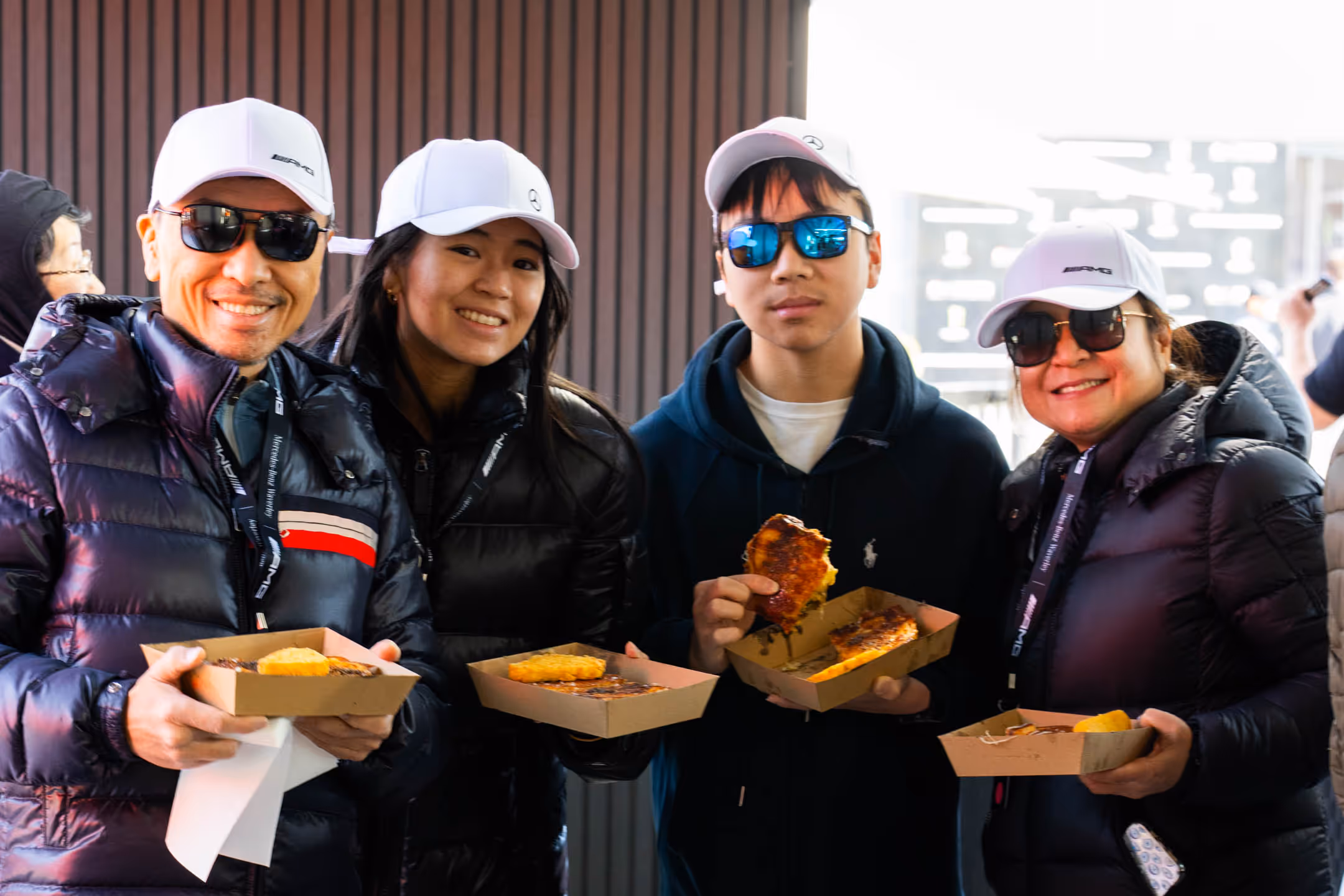 Four people in white caps and dark jackets holding trays with grilled sandwiches and yellow food items, smiling