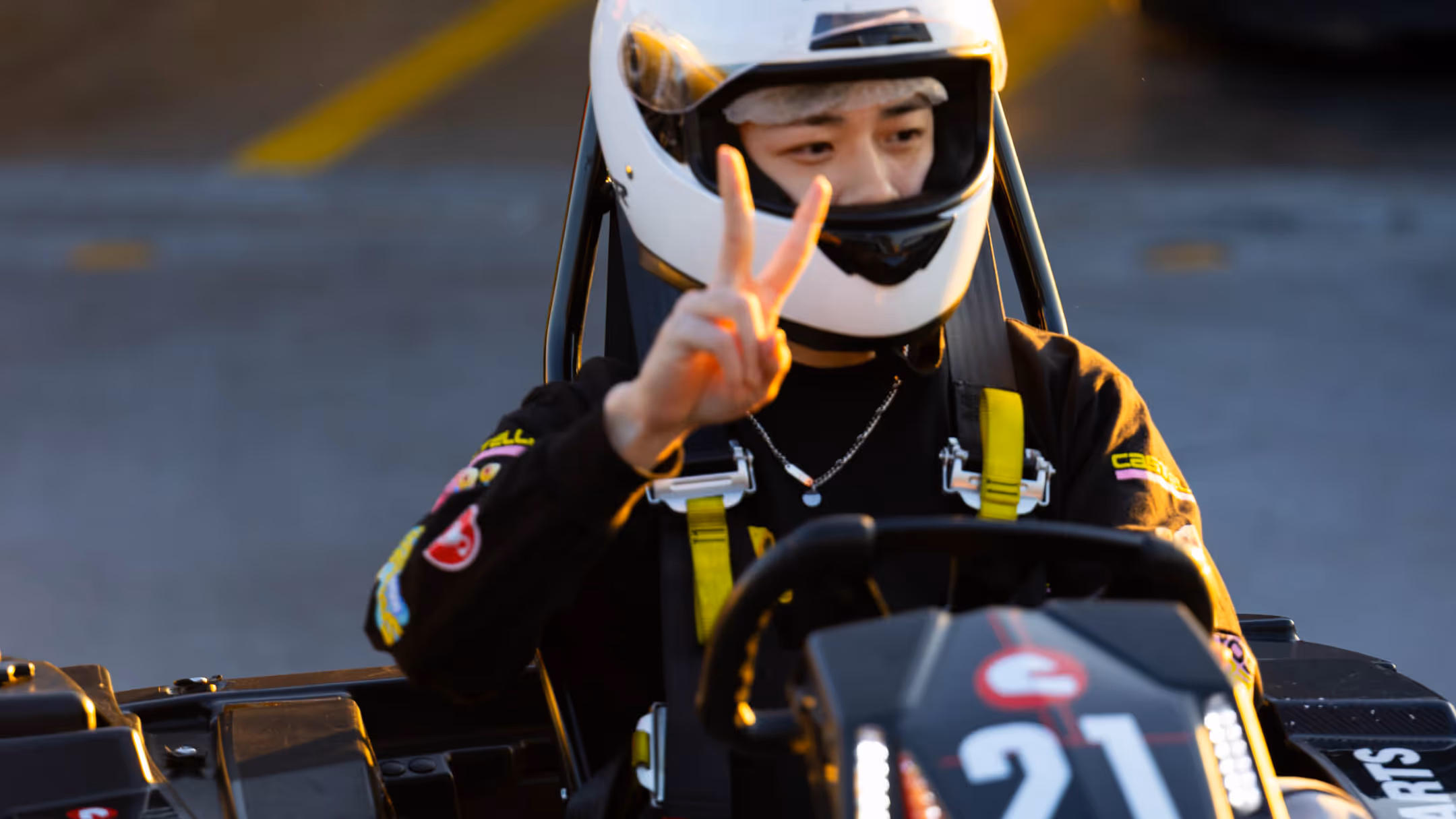 Person in white helmet seated in black go-kart with red accents making peace sign, yellow safety harnesses visible, number 21 on front