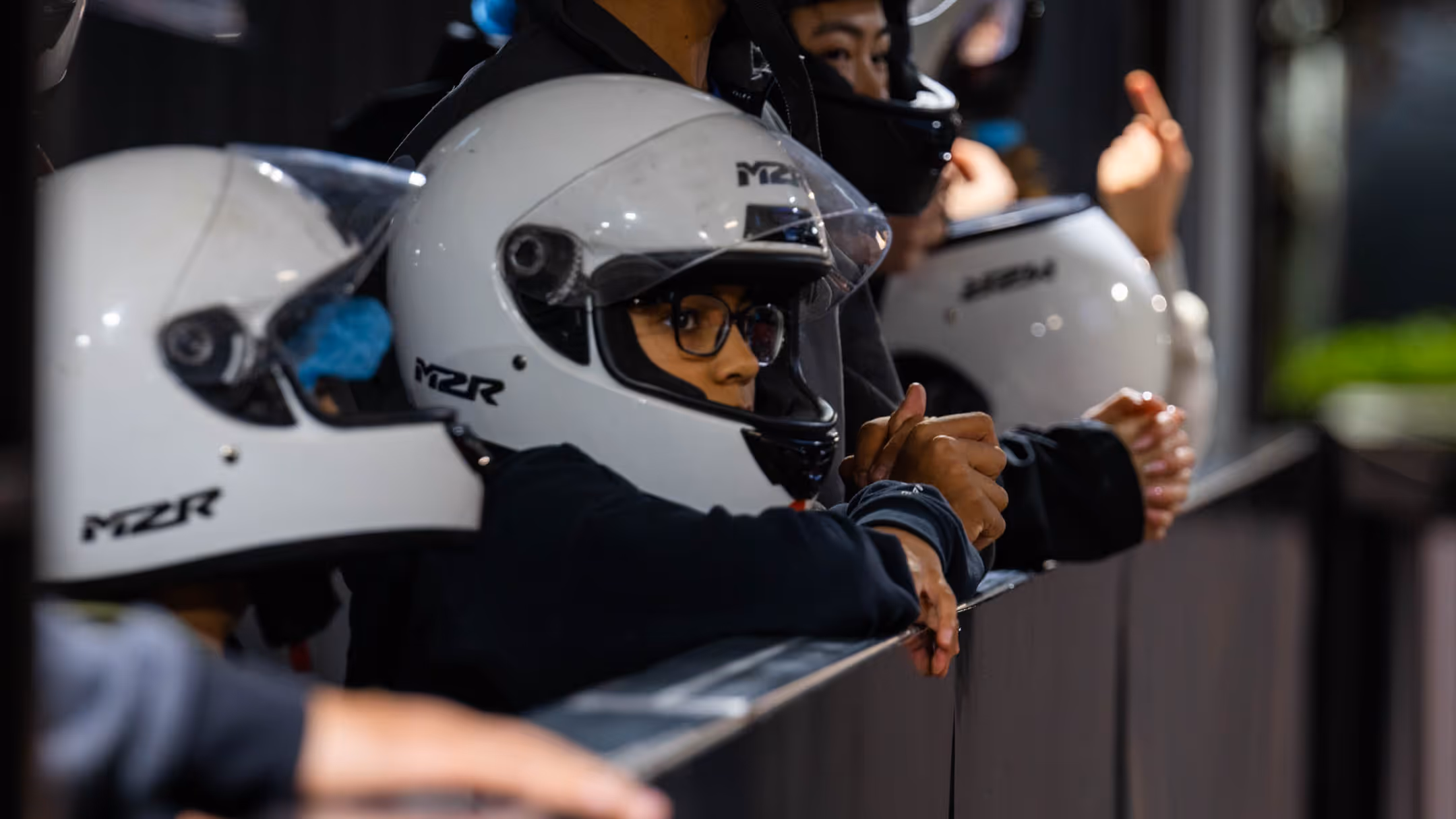 Group of people in white racing helmets with clear visors and MZR logos leaning on dark barrier, one person in glasses in foreground