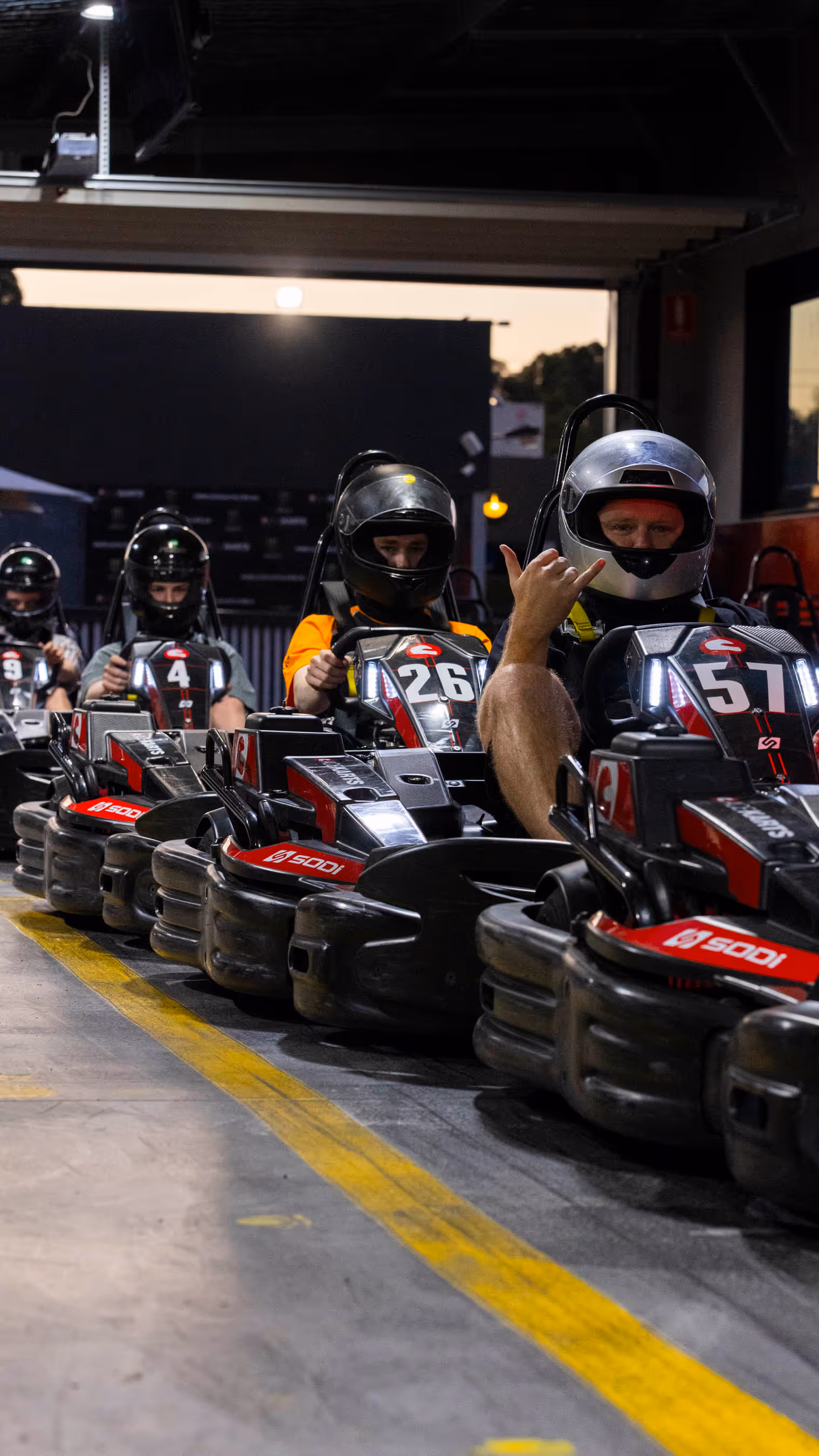 Line of five go-karts with drivers in helmets on concrete track, driver in kart 57 making shaka gesture, driver in kart 26 in orange shirt, yellow line on track