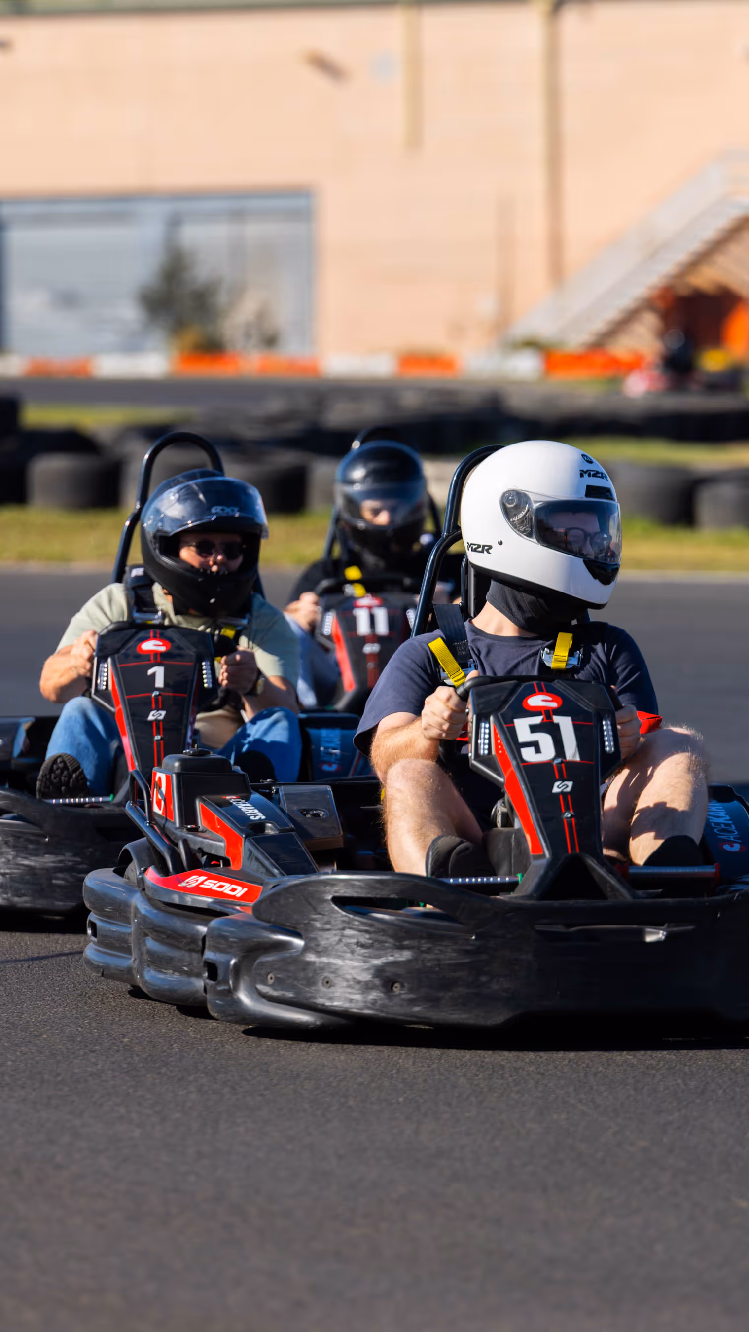 Three people in go-karts on asphalt track, white helmet driver in kart 51, black helmet drivers in karts 1 and 11, building and tire barriers in background