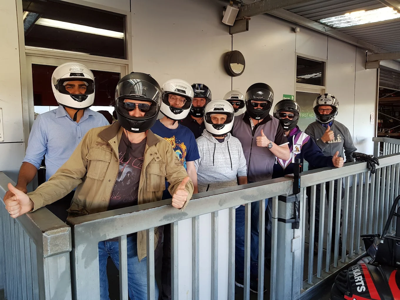 Group of people wearing helmets giving thumbs up at an outdoor go-kart track.