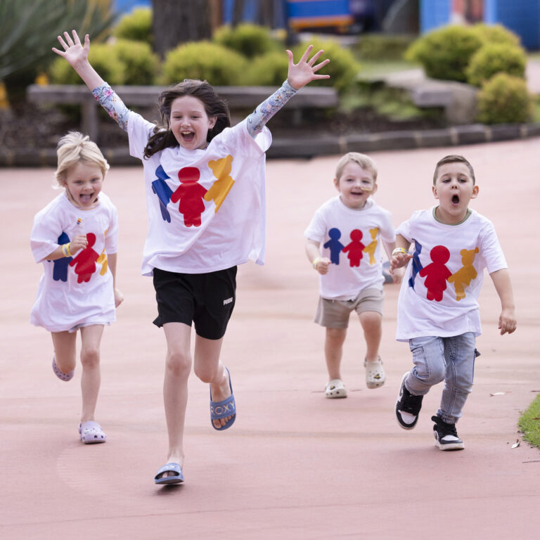 Four happy children wearing matching white graphic t-shirts run forward outdoors on a paved path with arms raised in joy.