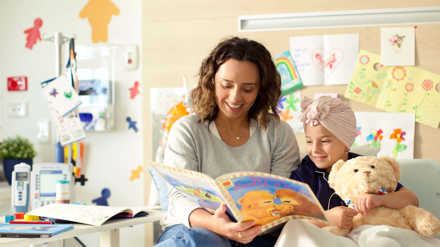 A woman reads a picture book to a young girl wearing a headwrap and holding a teddy bear in a colourful hospital room.