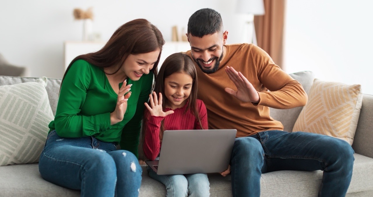 A family sitting on sofa. The mother, father and a young girl in the middle are waving to the laptop. 