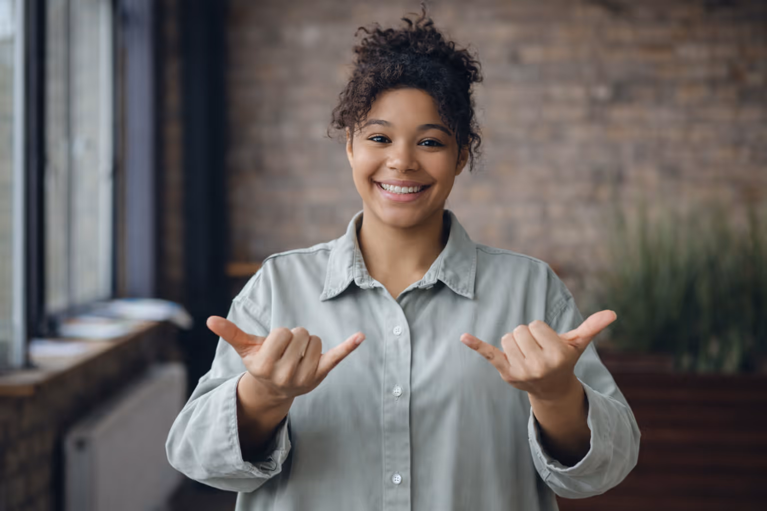 A light skinned brown female with light gray polo shirt signing now with brick background.