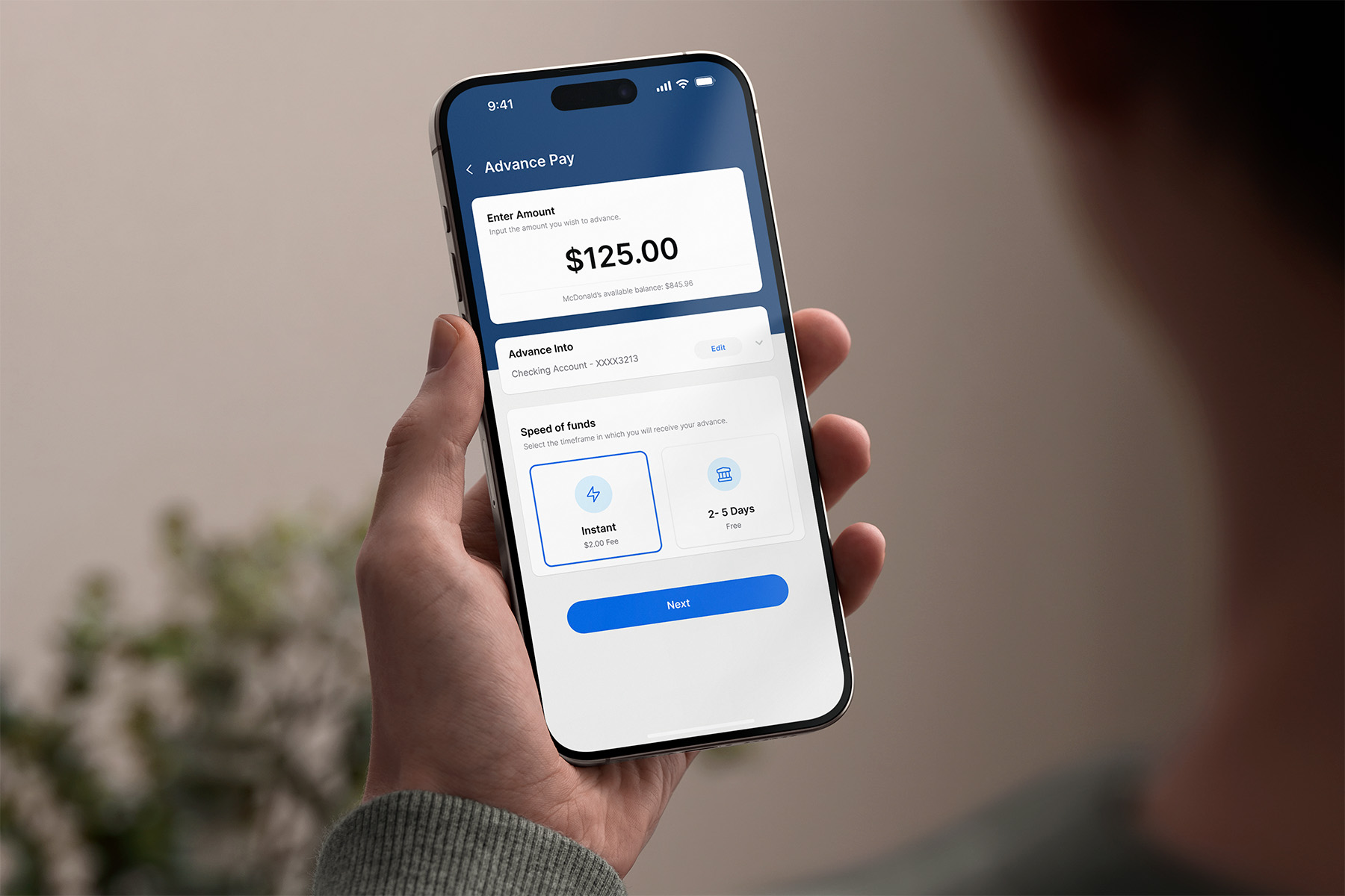 Hand holding a smartphone displaying an Advance Pay app screen with a $125.00 advance amount, checking account details, and options for instant or 2-5 days fund speed.