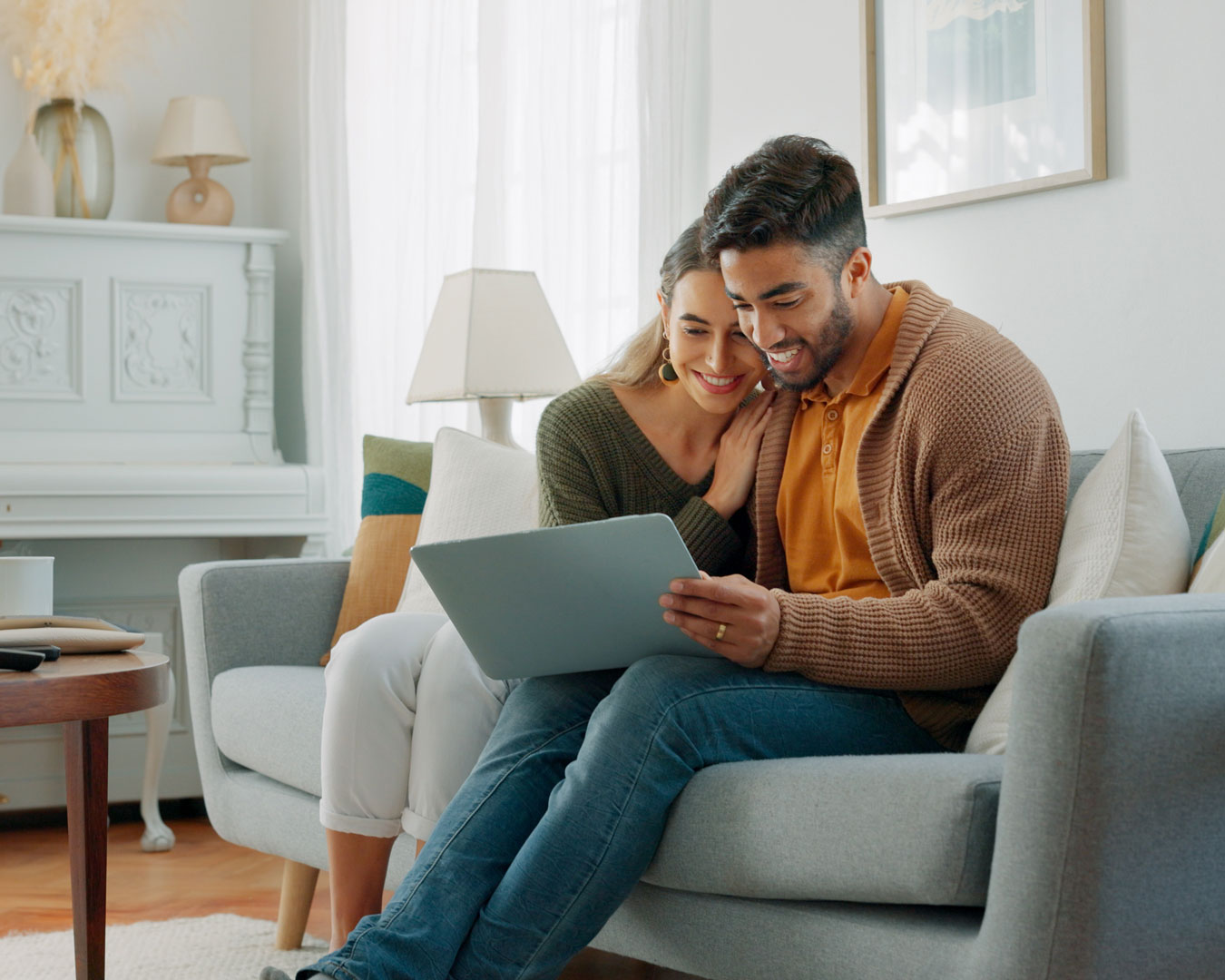 Smiling couple sitting on a gray sofa looking at a tablet together in a bright living room.