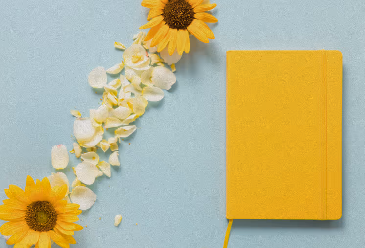 Un cahier jaune à élastique, à côté de deux tournesols et de pétales blancs éparpillés sur un fond bleu clair.