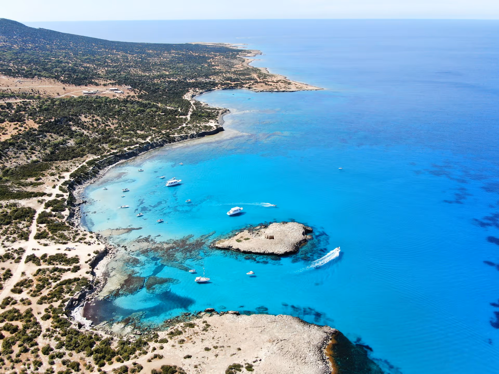 Aerial view of a coastal landscape with turquoise blue water, rocky shoreline, green vegetation, and several boats anchored near the shore.