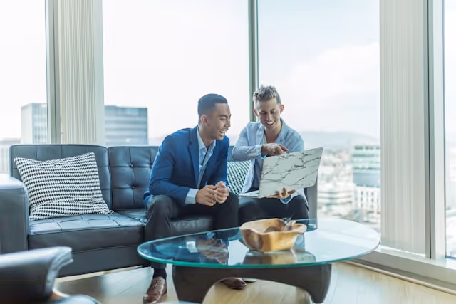 Two men in business attire sitting on a black couch and smiling while looking at a laptop in a modern office with large windows.