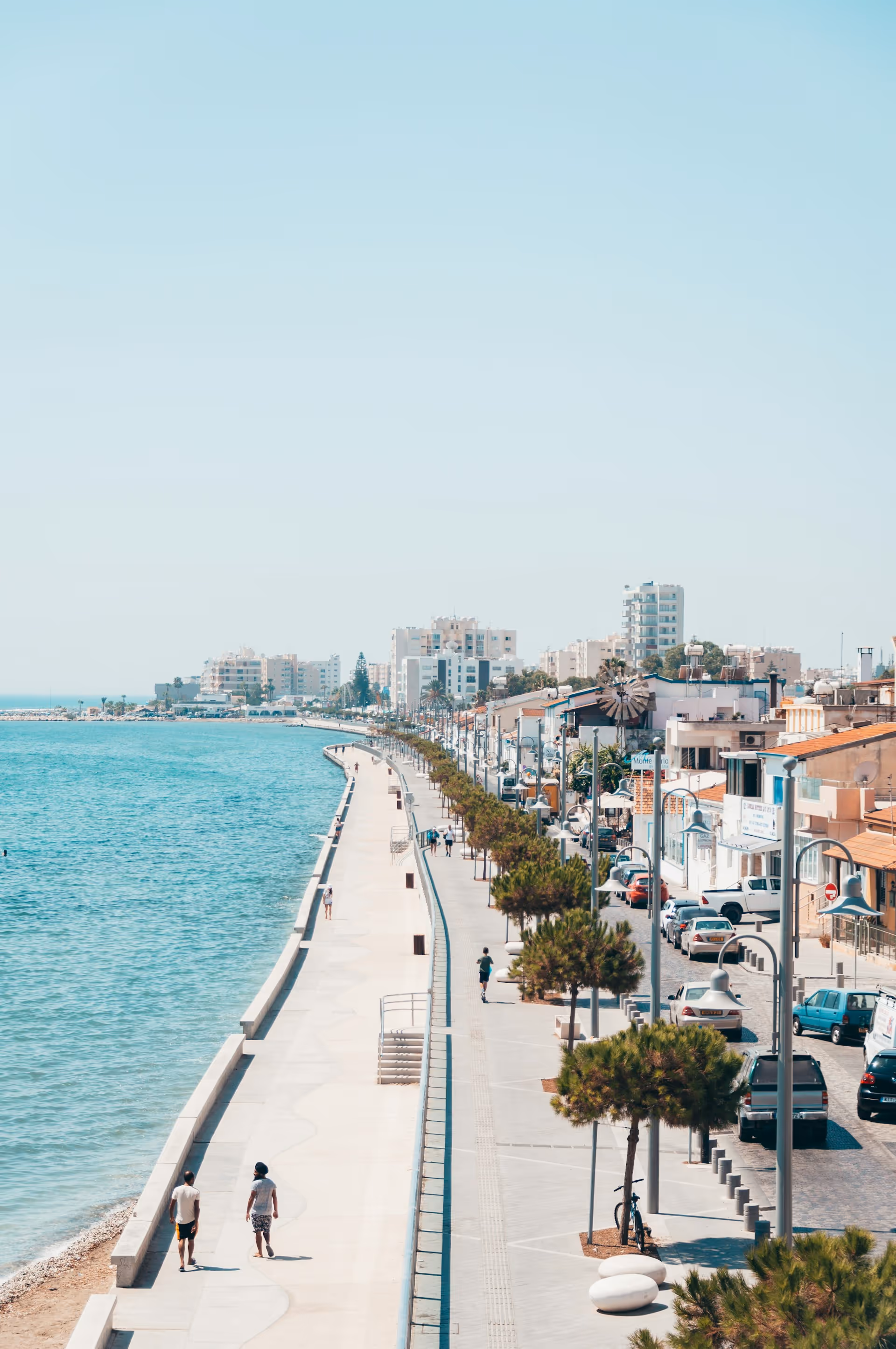 Coastal promenade with people walking, trees lining a street with parked cars, and buildings in the distance under a clear sky.