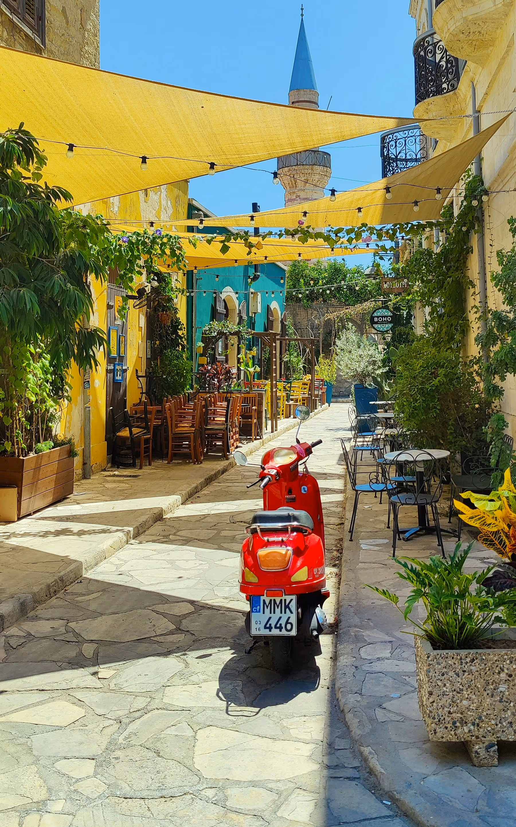 Red scooter parked on a sunny cobblestone street lined with outdoor café tables, greenery, and yellow awnings, with a minaret visible in the background.