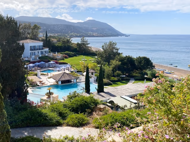Coastal resort with swimming pools, green lawns, trees, and hills in the background by the ocean under a partly cloudy sky.