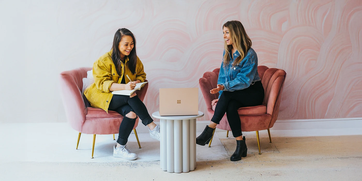 Two women sitting on pink chairs with a laptop on a small white table between them, engaged in conversation in a room with pink patterned wall.