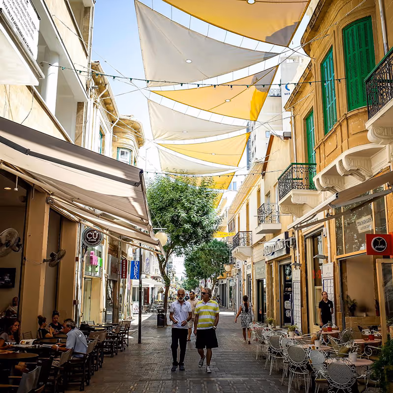 Sunny pedestrian street with cafes, outdoor seating, yellow and white shade sails overhead, and people walking.