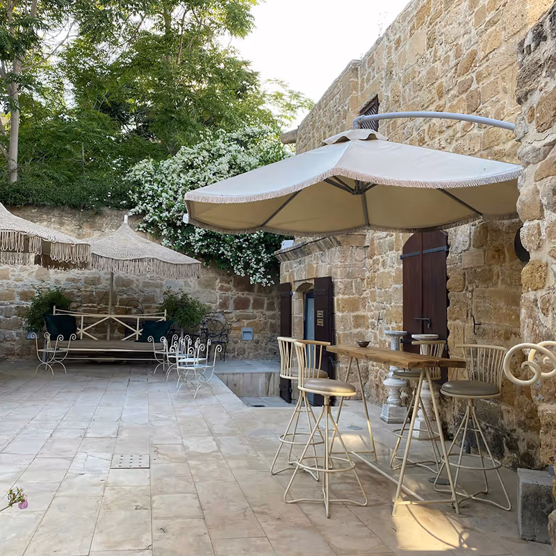 Outdoor patio with stone walls, a wooden high table with four metal bar stools under a large beige umbrella, and a swing with fringe umbrellas in the background.