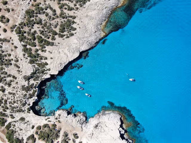 Aerial view of a rocky coastline with sparse vegetation and five boats floating in bright turquoise water.