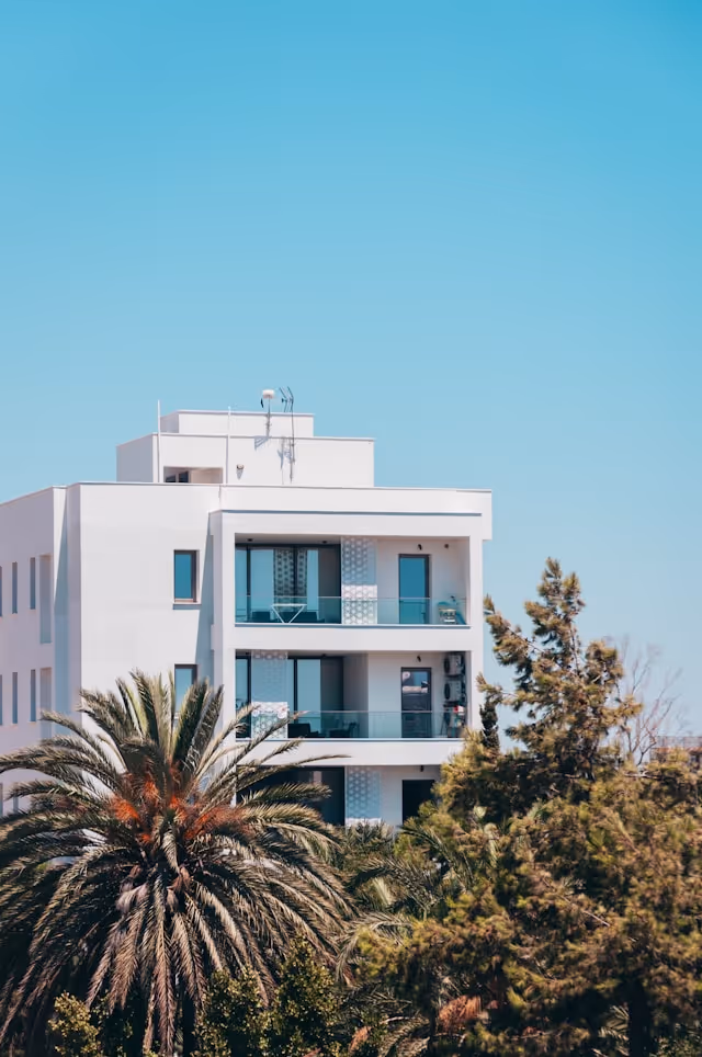 Modern white apartment building with balconies, surrounded by palm trees and greenery under a clear blue sky.