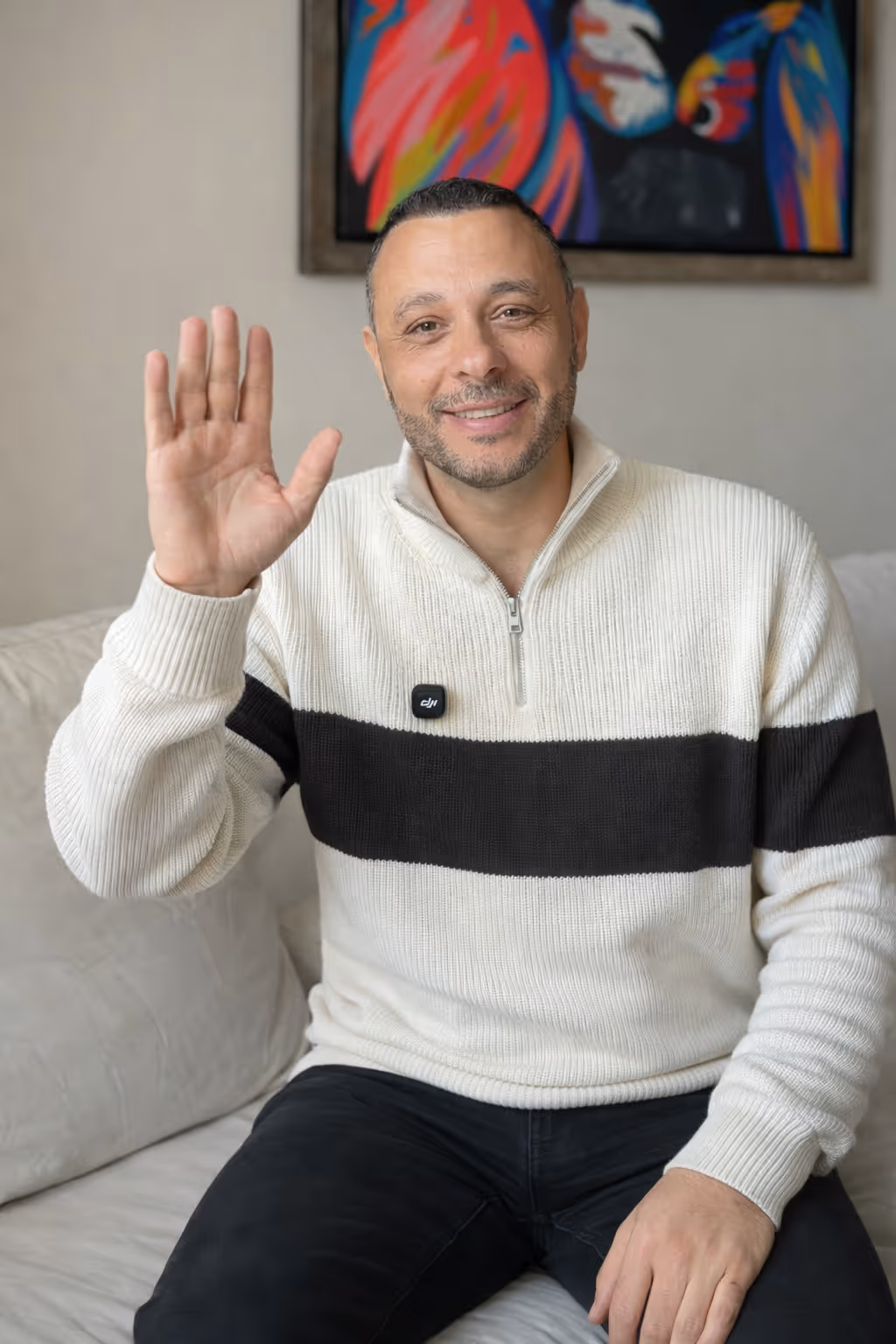 Smiling man with beard waving while sitting on a couch, wearing a white sweater with a black horizontal stripe.