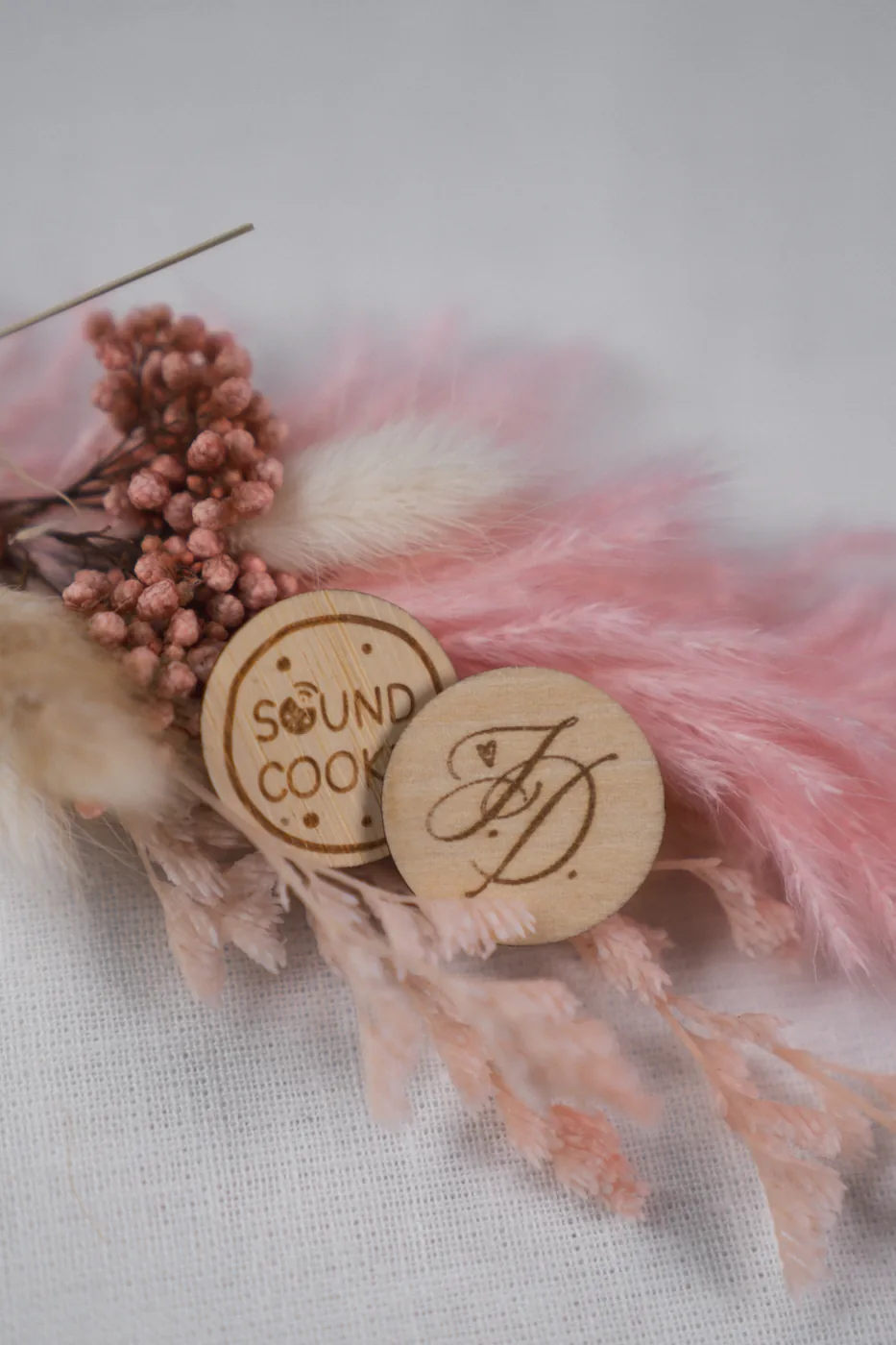 Zwei SoundCookies aus Holz, einer mit Monogramm graviert, zwischen rosafarbenen Trockenblumen.