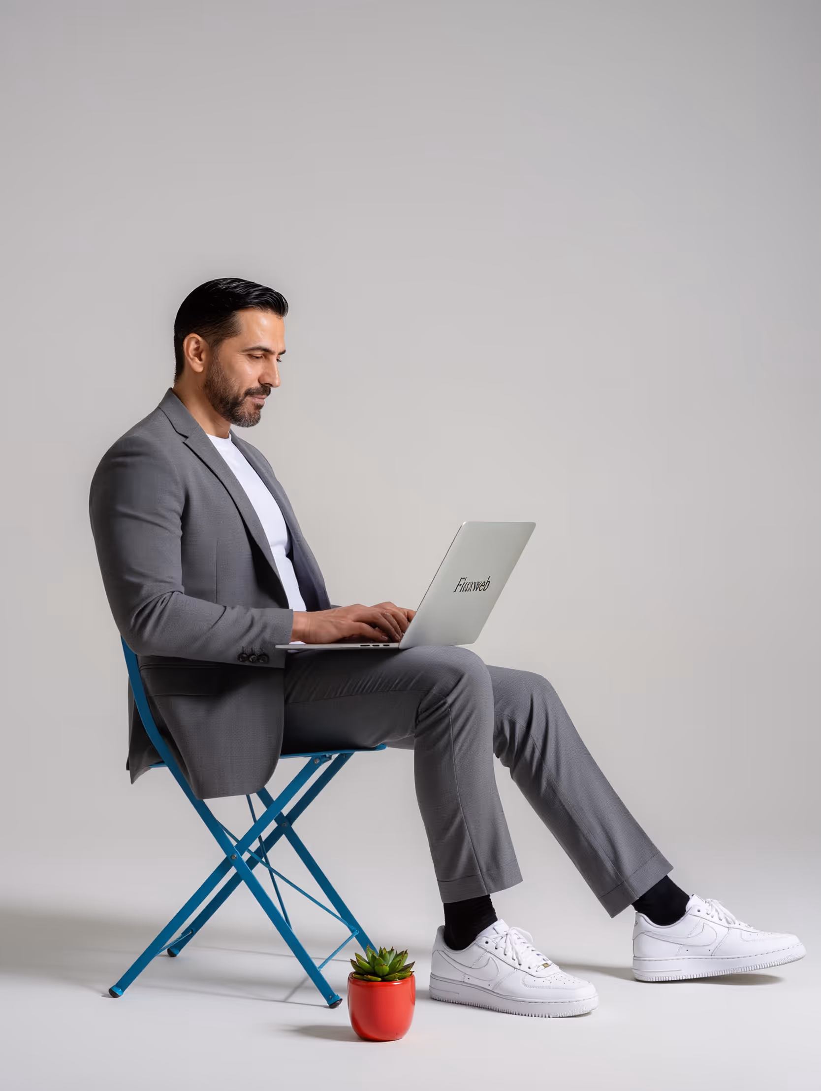 Homme en costume gris assis sur une chaise bleue travaillant sur un ordinateur portable avec plante en pot rouge.