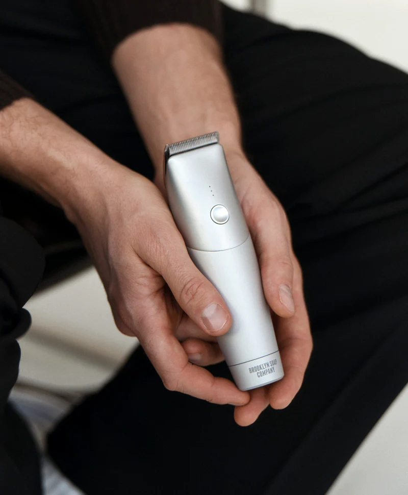 Person holding a silver electric hair trimmer with 'Brooklyn Soap Company' branding