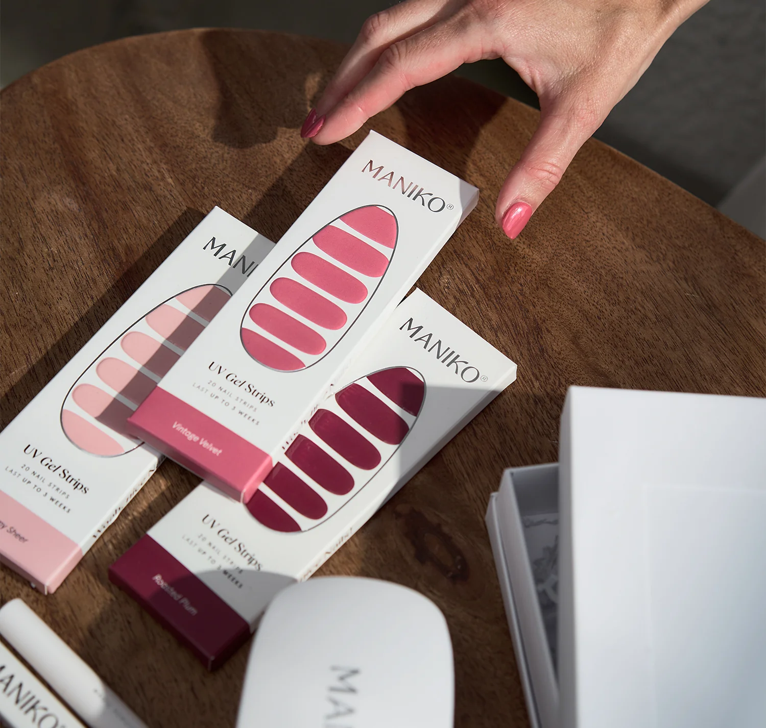 Hand with pink manicured nails reaching toward Maniko UV gel nail strips in pink and burgundy shades on a wooden surface