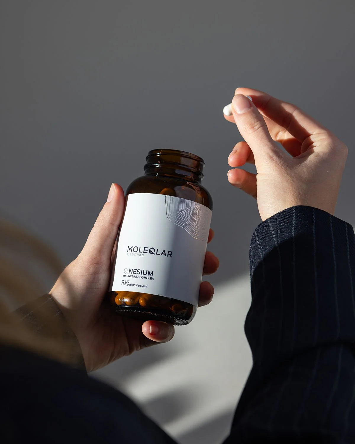 Person holding a dark glass bottle labeled Molecular Essentials Qnesium Magnesium Complex and a white capsule between fingers.
