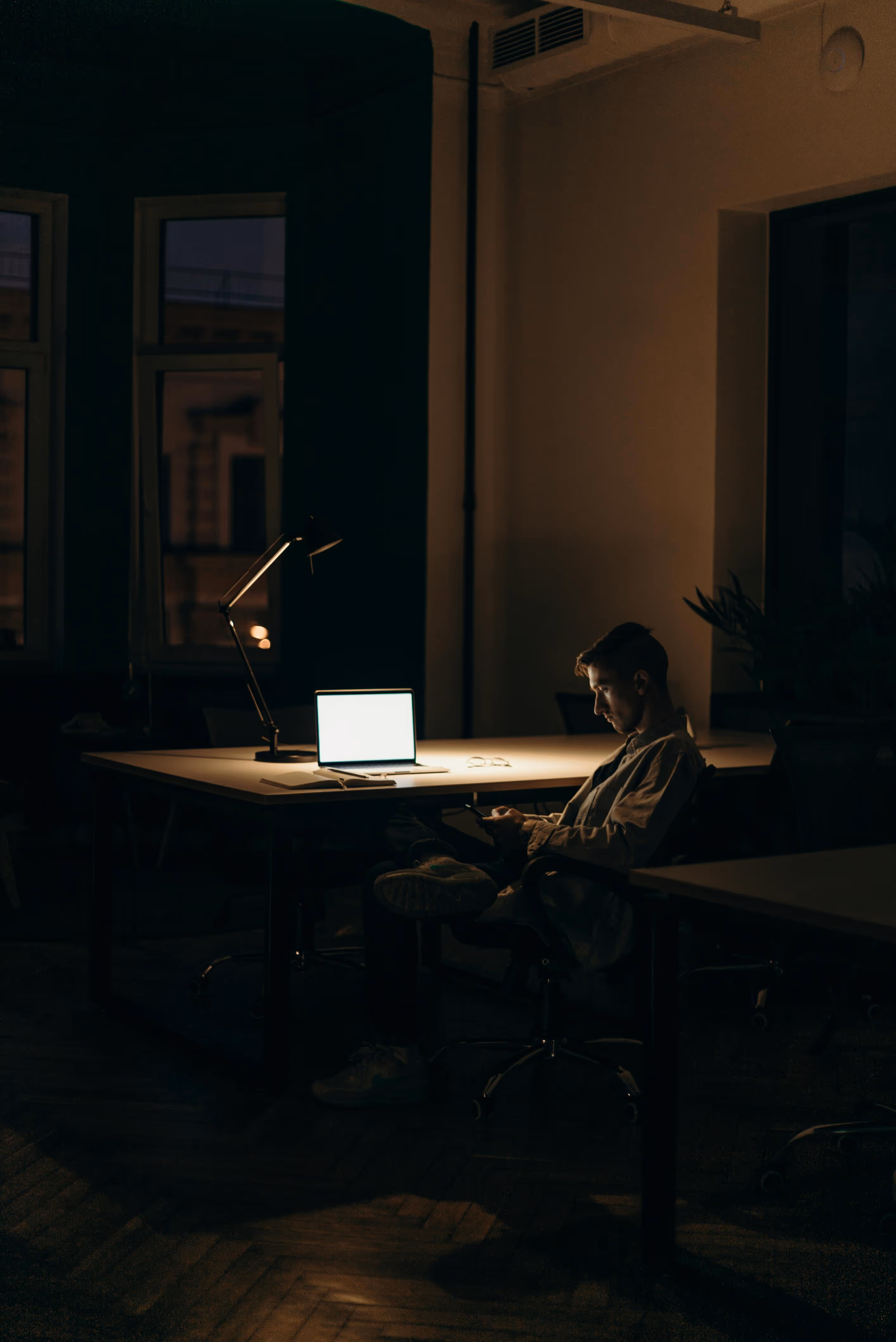 Junger Mann sitzt abends in einem dunklen Büro und schaut auf sein Smartphone neben einem beleuchteten Laptop.