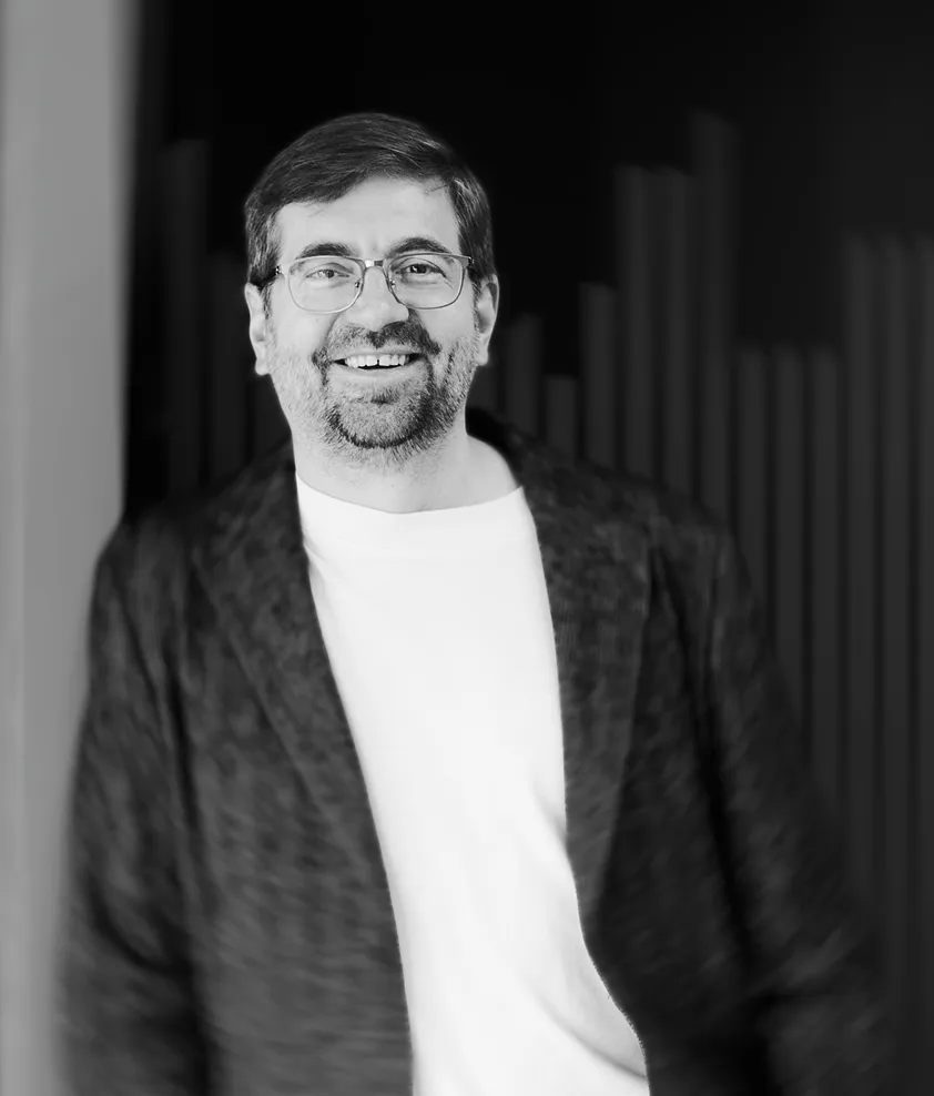 Smiling man with glasses, beard, and mustache wearing a white shirt and dark jacket against a dark background.