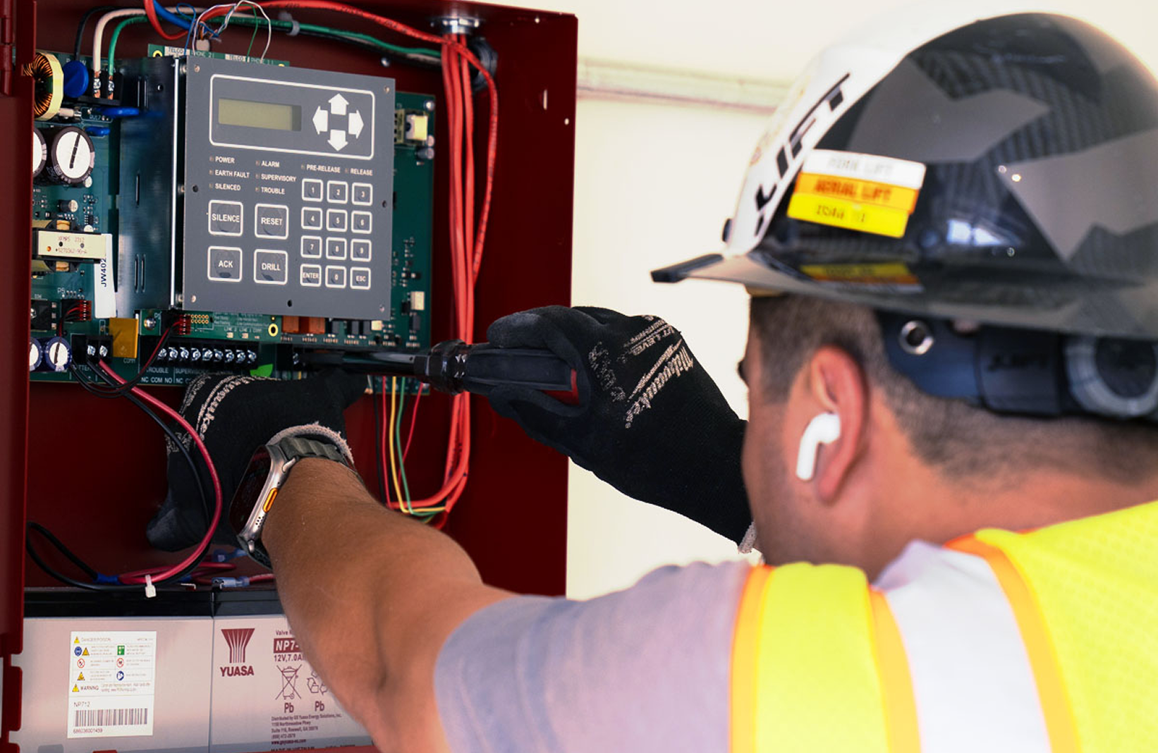 Technician wearing gloves and helmet using screwdriver to work on electrical control panel.
