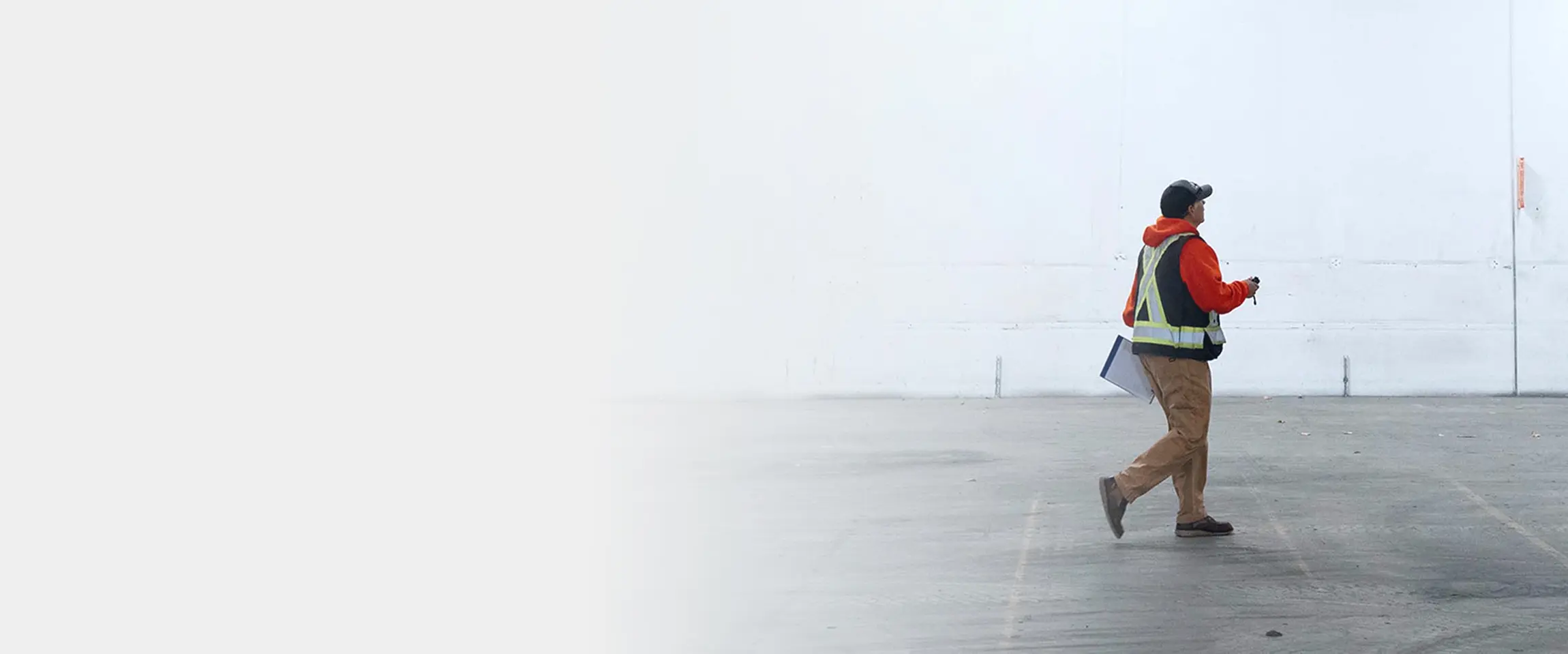 Worker in safety vest and hat walking inside a large empty warehouse holding a clipboard and device.