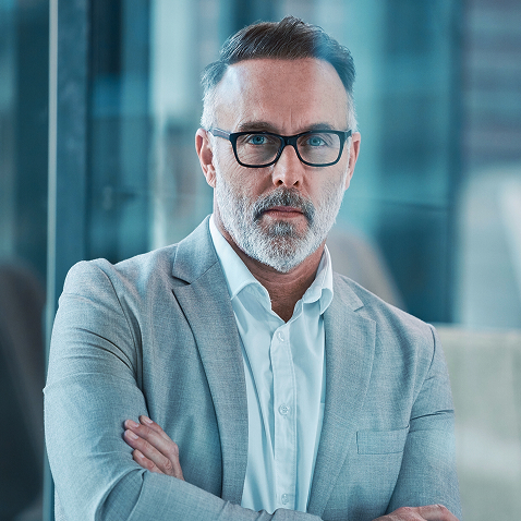 Middle-aged man with gray hair and beard wearing glasses and a gray suit, arms crossed.