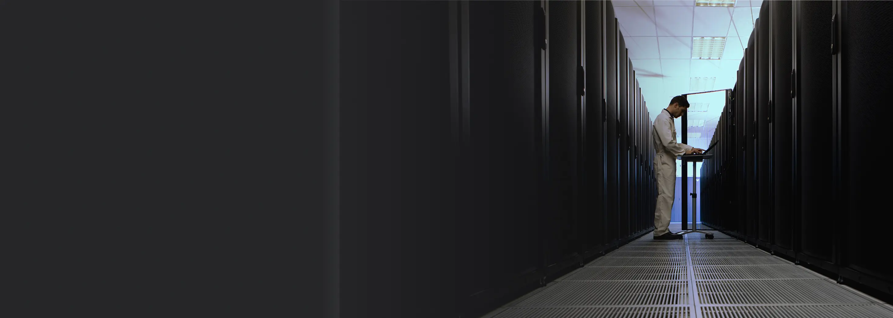 IT technician working on a laptop in a server room with racks on both sides.