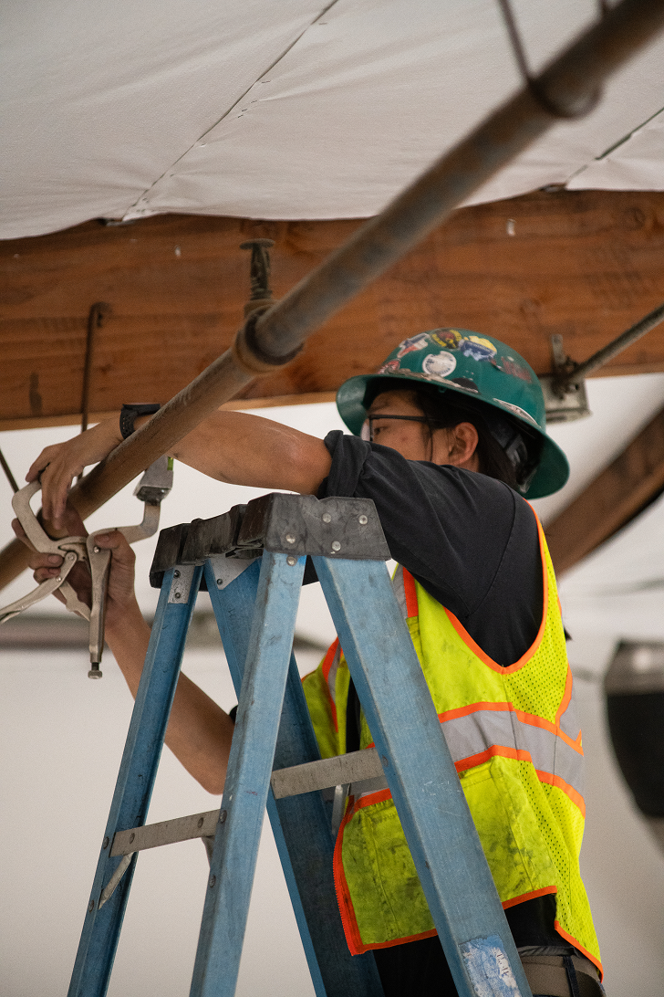 Construction worker wearing a green helmet and yellow safety vest uses a tool on overhead pipe from a ladder.