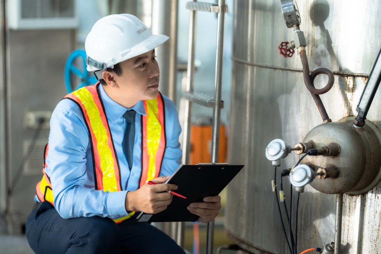 Engineer in safety vest and helmet inspecting industrial tank with clipboard and pen.